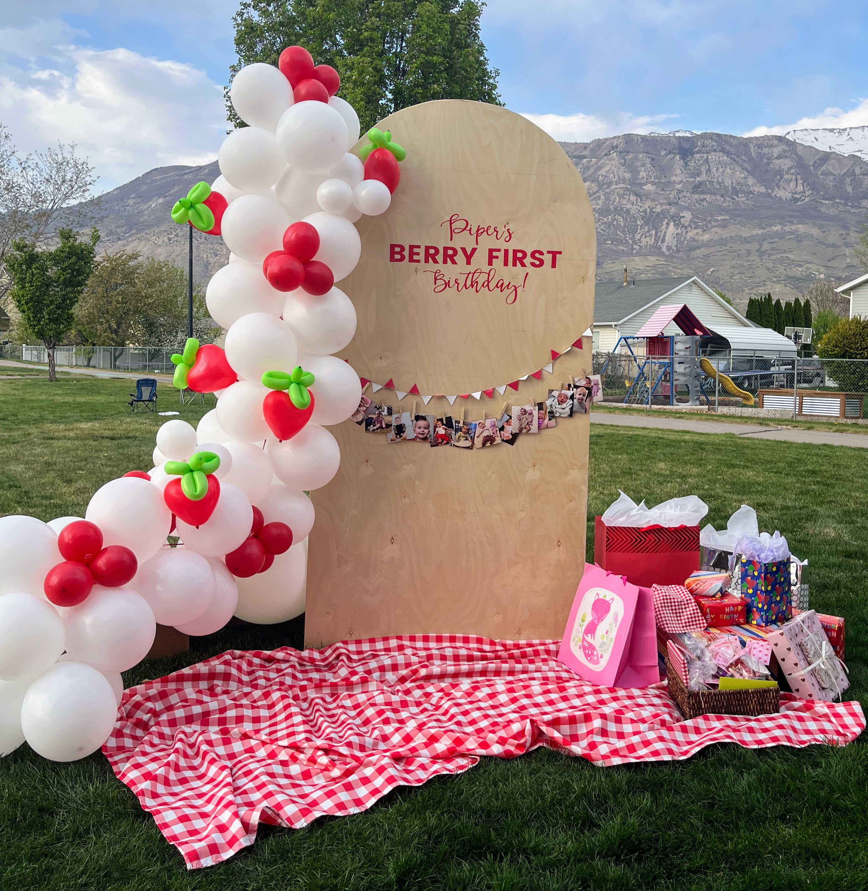 Decorative setup for a berry-themed first birthday party with balloons, a cardboard arch, and gifts on a grassy field.