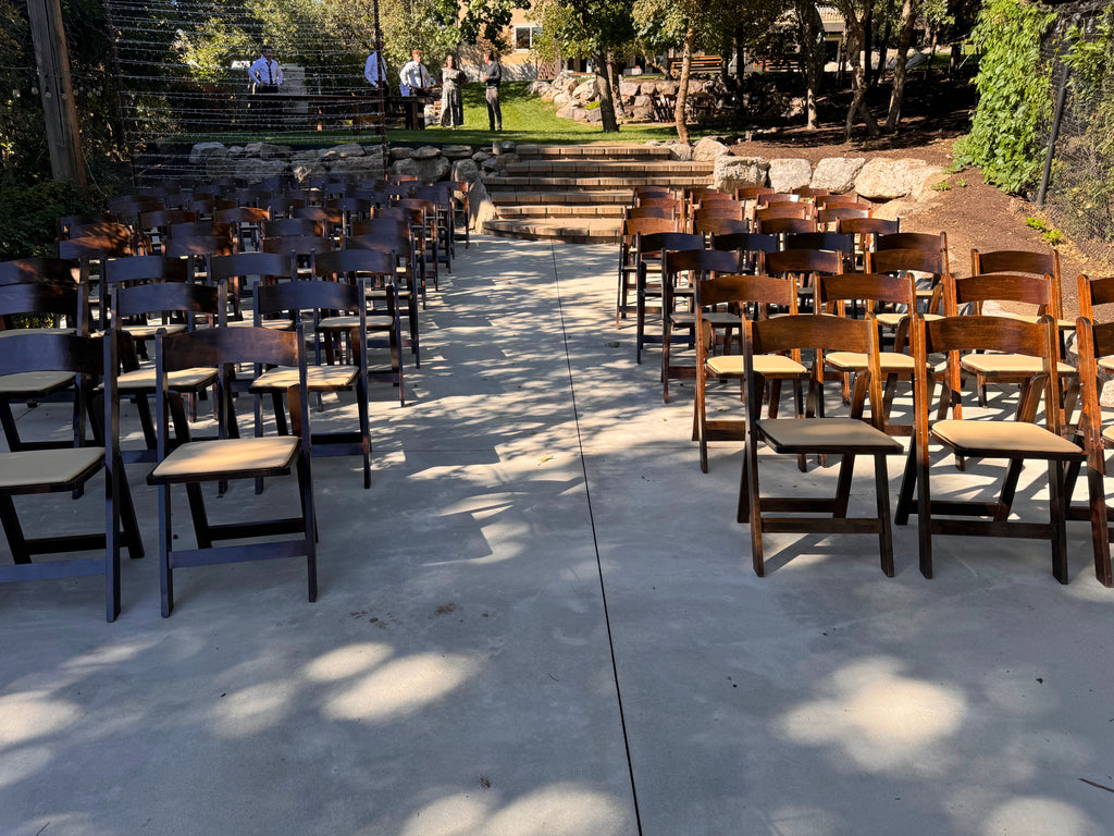 Rows of wooden chairs set up on a concrete surface outdoors with trees in the background.