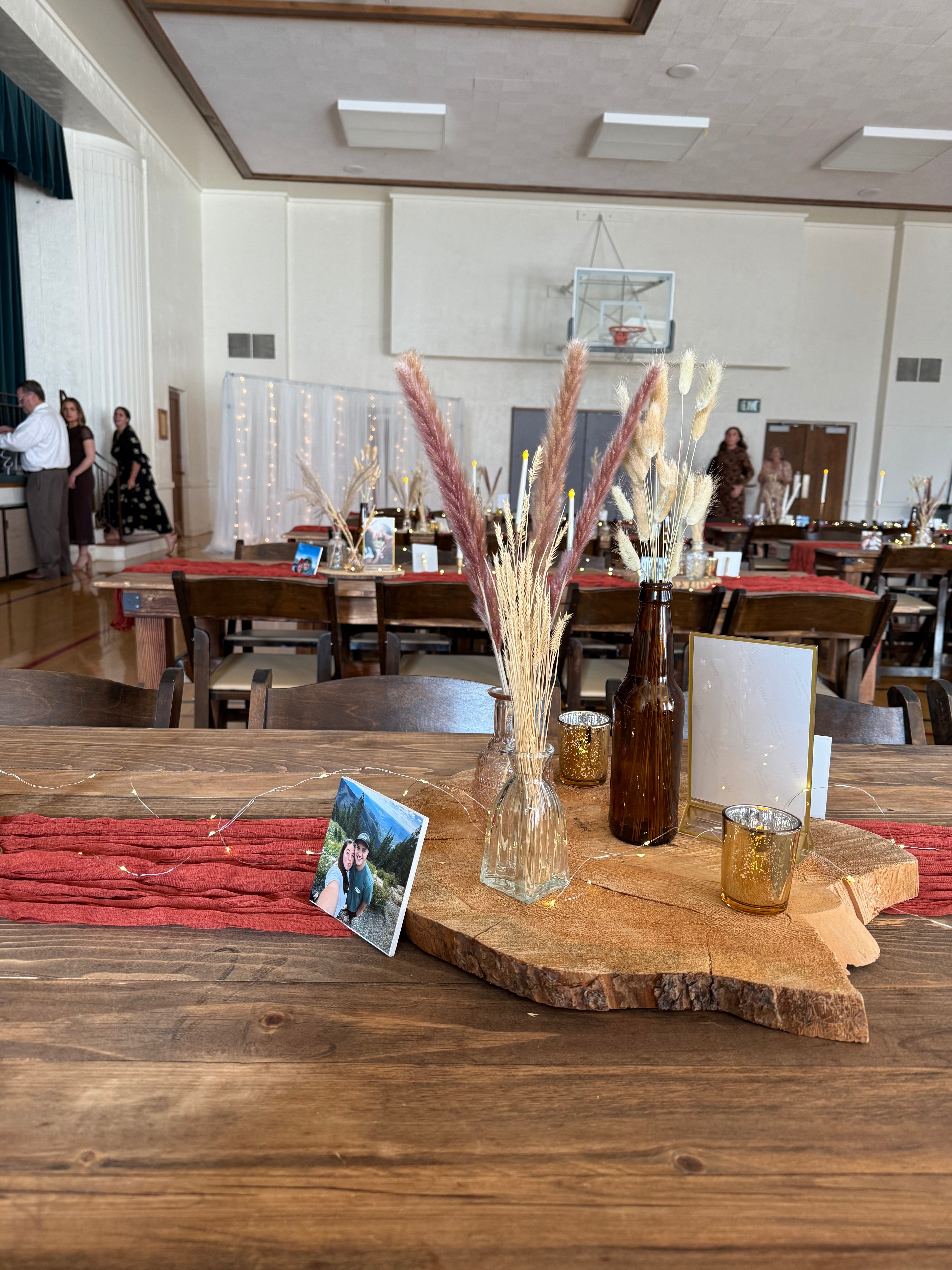 Decorated table with a vase of dried grasses, bottle, and glass on a wooden table in a large room.