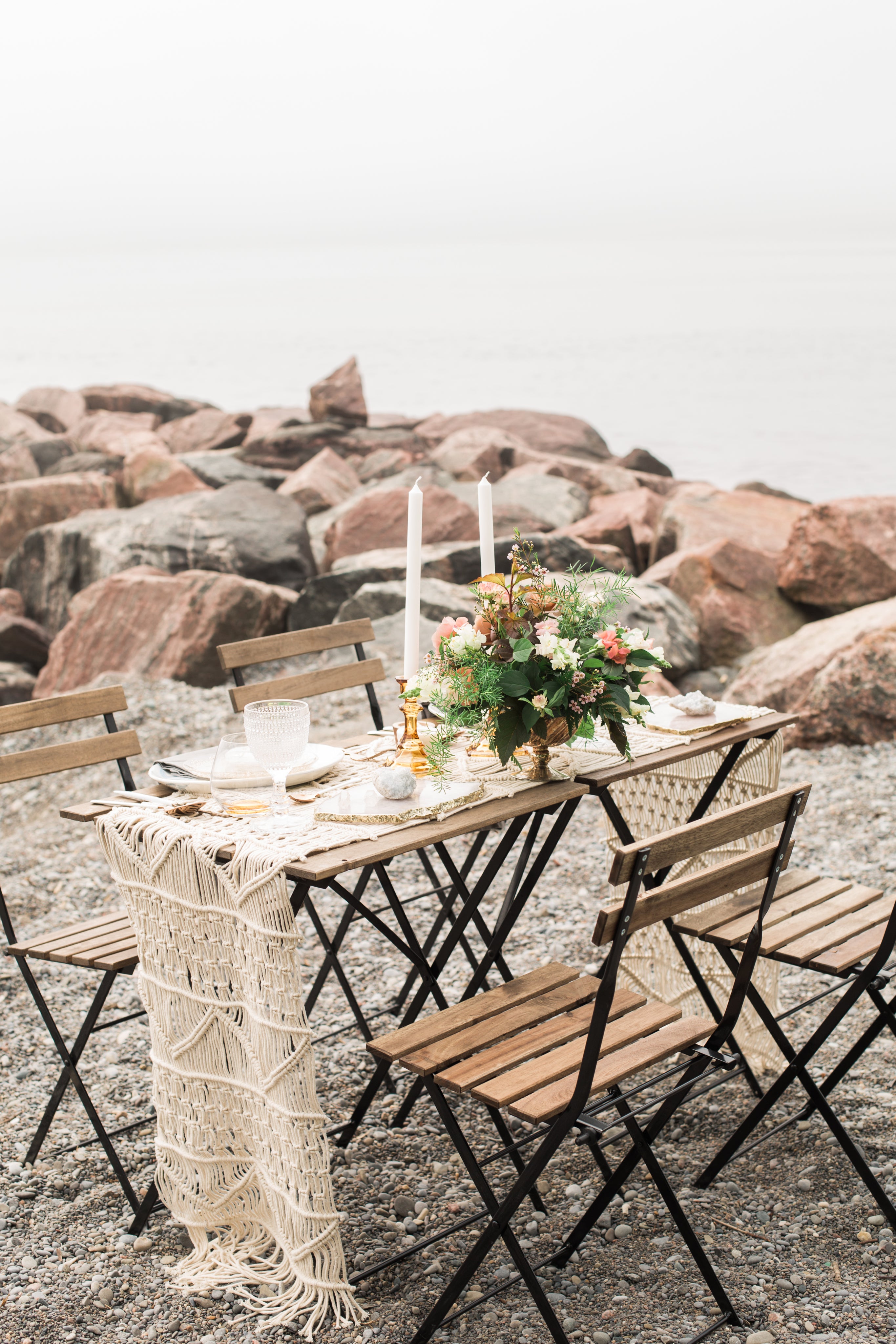 table with candles, flowers and dishes along with chairs set on a beach with rocks