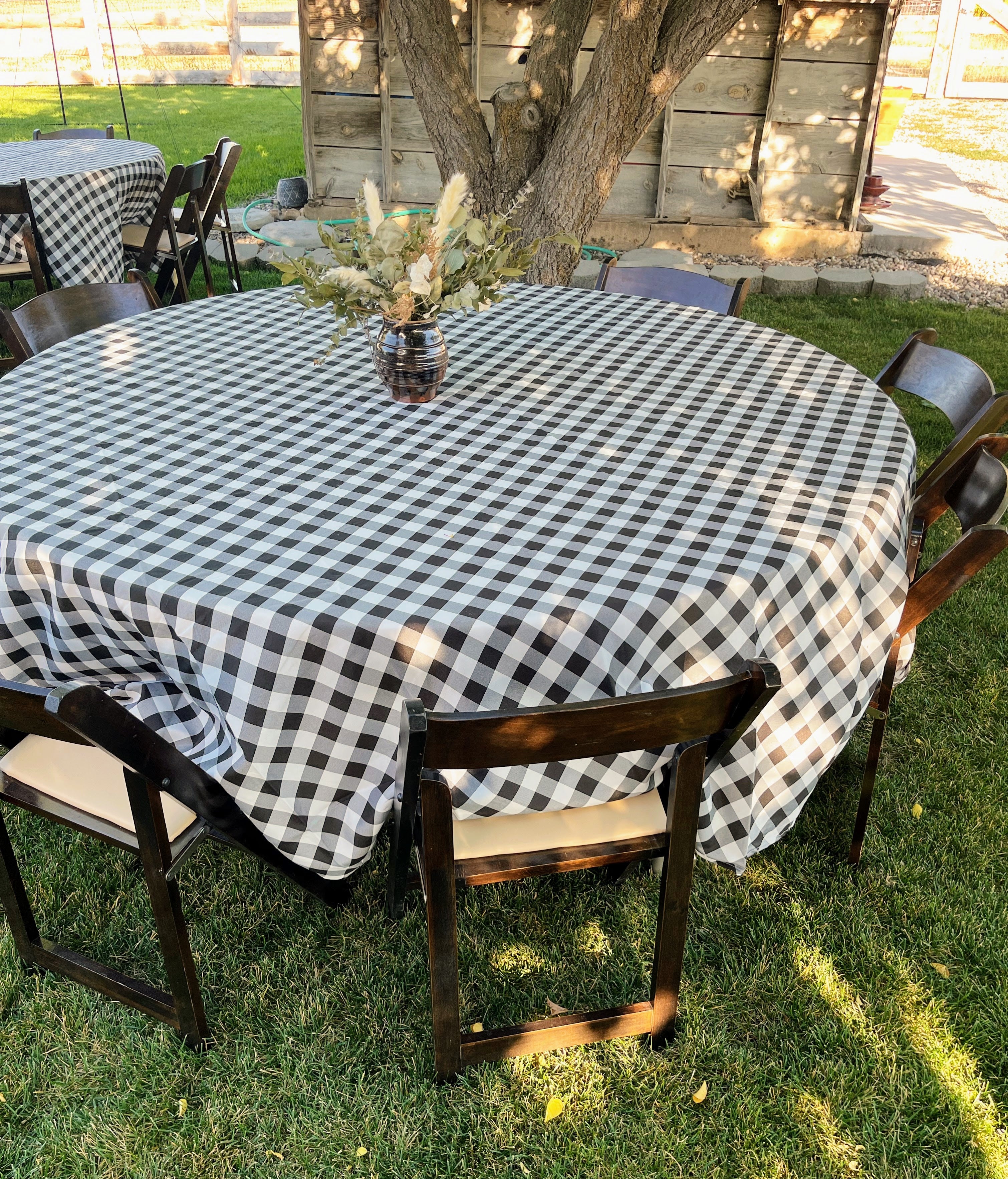 Round table with a black and white checkered tablecloth surrounded by chairs on a grassy outdoor area.